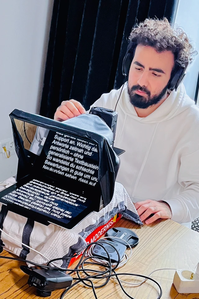 Auf einem Foto ist ein Mann zu sehen, der an einem Holztisch sitzt, einen weißen Kapuzenpulli und schwarze Kopfhörer trägt. Er hat lockiges Haar und einen Bart und ist auf einen vor ihm aufgestellten Teleprompter konzentriert. Der Bildschirm des Teleprompters zeigt weißen Text auf schwarzem Hintergrund, der teilweise sichtbar ist. Der Teleprompter ist auf einem Bücherstapel platziert und mit einem Tuch abgedeckt. Auf dem Tisch liegen verschiedene Kabel und ein Kameraobjektivdeckel. Im Hintergrund befindet sich ein dunkler Vorhang, der den Kontrast zu dem gut beleuchteten Arbeitsbereich verstärkt.