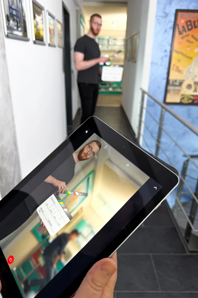 Auf einem Foto ist eine Person zu sehen, die ein Tablet in der Hand hält, auf dem das Bild eines Mannes mit einer Filmklappe zu sehen ist. Der Mann auf dem Bildschirm des Tablets hat rote Haare und eine Brille und steht in einem Flur. Der echte Mann, ebenfalls mit roten Haaren und Brille, ist im Hintergrund des Fotos zu sehen, leicht unscharf, und steht im selben Korridor. Der Flur weist weiße Wände mit gerahmten Bildern und eine blaue Wand mit einem bunten Poster auf. Die Szene suggeriert einen Blick hinter die Kulissen eines Dreh- oder Produktionsprozesses, in dem sich digitale und reale Elemente vermischen.
