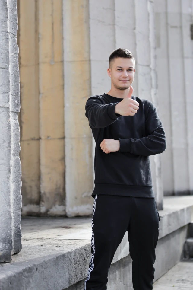 Eine Fotografie eines jungen Mannes, der im Freien vor einer Reihe hoher Steinsäulen steht. Er ist mit einem schwarzen Sweatshirt und einer schwarzen Hose mit weißen Streifen an den Seiten bekleidet. Der Mann lächelt und zeigt mit der rechten Hand den Daumen nach oben, während er den linken Arm lässig vor der Brust verschränkt. Der Hintergrund zeigt die Textur und die Farbvariationen der Steinsäulen, was auf eine architektonische oder historische Umgebung hinweist. Die Beleuchtung ist natürlich, was darauf hindeutet, dass das Foto am Tag aufgenommen wurde.