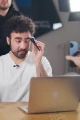 A photograph captures a young man with curly hair and a beard, wearing a white shirt, seated at a table with a laptop in front of him. He is holding a makeup brush near his face, appearing to apply or remove makeup from his right eye. Another person is partially visible in the background, blurred, and a hand holding a smartphone is visible in the foreground, suggesting that the scene is being recorded or live-streamed. The setting appears to be a studio with professional lighting equipment visible in the background, creating a focused and well-lit environment.