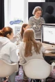 A photograph captures a group of young women working together in a bright, well-lit room. In the foreground, three women are seated at a table, focused on a large computer monitor displaying a design or text. They are wearing casual clothing, including hoodies. In the background, a woman with short blonde hair and glasses, dressed in a striped sweater, stands smiling with her arms crossed, overseeing the activity. The setting appears to be a collaborative workspace with a large window letting in natural light and a decorative rug on the floor. Various signs and materials are visible in the background, suggesting a creative and engaging environment.