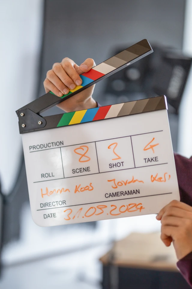 The image shows a close-up of a person holding a clapperboard (also known as a slate) used in film production. The clapperboard has a colorful top bar with black, white, red, yellow, green, and blue stripes. The bottom section is divided into sections with text written in orange marker. A hand is holding the top bar, preparing to snap it shut. The background is blurred, indicating an indoor setting, likely on a film set. The clapperboard is used to mark scenes and takes during filming.