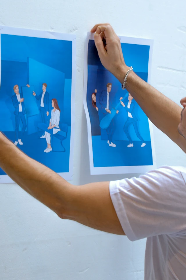 Eine Fotografie zeigt eine Person, die zwei Bildabzüge an eine weiße Wand pinnt. Die Person, die ein weißes T-Shirt und ein silbernes Armband trägt, ist von der Seite zu sehen und konzentriert sich auf ihre Aufgabe. Die Drucke zeigen leuchtend blaue Hintergründe mit Illustrationen oder Fotos von Personen in blauer Kleidung. Auf einem Bild sind drei Personen zu sehen, von denen eine sitzt und die anderen beiden stehen und sich unterhalten. Das zweite Bild zeigt zwei Personen in einer dynamischen Pose, wobei eine dritte Person teilweise sichtbar ist. Die Gesamtszene deutet auf einen künstlerischen oder kreativen Arbeitsbereich hin, in dem die Person die Drucke für die Ausstellung oder Überprüfung sorgfältig arrangiert.