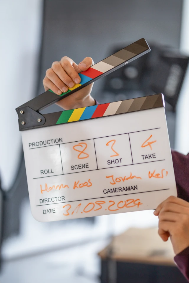Das Bild zeigt eine Nahaufnahme einer Person, die eine in der Filmproduktion verwendete Klappe (auch Schiefertafel genannt) hält. Die Klappe hat eine bunte obere Leiste mit schwarzen, weißen, roten, gelben, grünen und blauen Streifen. Der untere Teil ist in Abschnitte unterteilt, die mit orangefarbenen Markern beschrieben sind. Eine Hand hält die obere Leiste und bereitet sich darauf vor, sie zuzuschlagen. Der Hintergrund ist unscharf, was auf einen Innenraum hindeutet, wahrscheinlich auf ein Filmset. Die Klappe wird verwendet, um Szenen und Takes während der Dreharbeiten zu markieren.