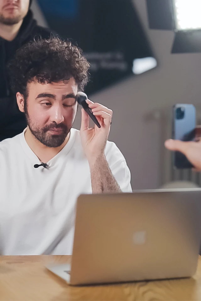 Das Foto zeigt einen jungen Mann mit lockigem Haar und Bart, der ein weißes Hemd trägt und an einem Tisch sitzt, vor dem ein Laptop steht. Er hält einen Schminkpinsel in der Nähe seines Gesichts und scheint sein rechtes Auge zu schminken oder zu entfernen. Eine weitere Person ist teilweise im Hintergrund zu sehen, unscharf, und eine Hand, die ein Smartphone hält, ist im Vordergrund zu sehen, was darauf hindeutet, dass die Szene aufgezeichnet oder live gestreamt wird. Der Schauplatz scheint ein Studio zu sein, in dem im Hintergrund eine professionelle Beleuchtungsanlage zu sehen ist, die eine konzentrierte und gut beleuchtete Umgebung schafft.