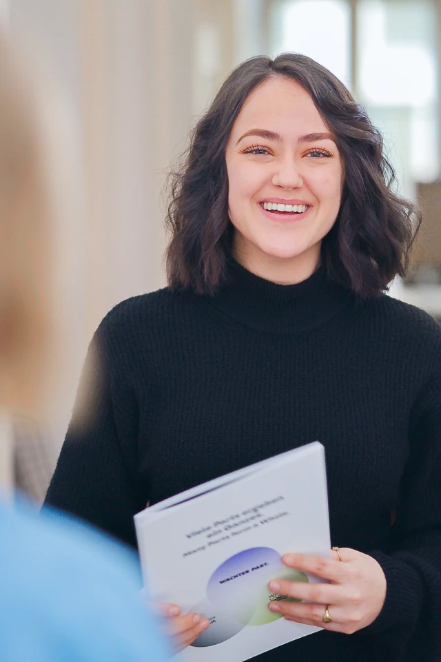 A photograph captures a young woman with shoulder-length dark hair, smiling brightly. She is wearing a black turtleneck sweater and holding a booklet or folder with both hands. The booklet has a design on the cover with some text and a circular graphic. The background is softly blurred, indicating an indoor setting with natural light coming from a window or doorway. The overall mood of the image is warm and welcoming, highlighting the woman’s cheerful expression.