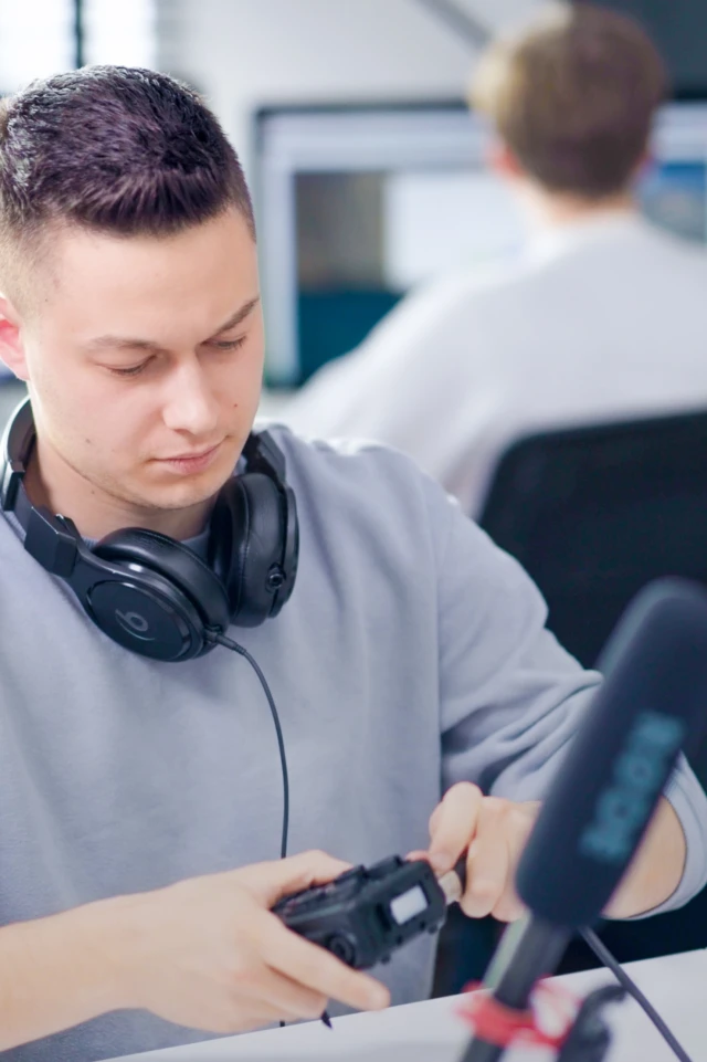 Ein Foto zeigt einen jungen Mann mit kurzen dunklen Haaren, der ein hellgraues Sweatshirt trägt und an einem Schreibtisch sitzt. Er hat ein Paar große schwarze Kopfhörer um den Hals und ist damit beschäftigt, ein Gerät in seinen Händen einzustellen, möglicherweise eine Kamera oder ein Audiogerät. Vor ihm ist ein Mikrofon auf einem Ständer zu sehen. Der Hintergrund ist unscharf und zeigt eine weitere Person, die an einem Computer arbeitet, was auf ein Büro oder ein Studio hindeutet. Die gesamte Szene deutet auf eine professionelle oder kreative Arbeitsumgebung hin.