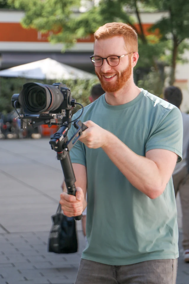 A photograph shows a young man with red hair and a beard, wearing glasses and a light green t-shirt, operating a professional camera mounted on a stabilizing gimbal. He is smiling as he focuses on capturing footage. The scene takes place outdoors in a well-lit area, with blurred trees and buildings in the background, suggesting an urban setting. The man appears to be enjoying his task, and the setup indicates a professional or semi-professional video recording activity.