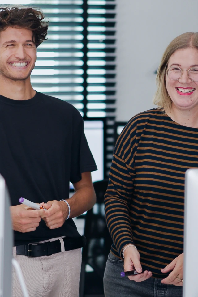 A photograph shows two young adults standing and smiling in a brightly lit indoor setting. The man on the left has curly hair and is wearing a black t-shirt and light-colored pants with a black belt. He is holding a marker in his hands. The woman on the right has blonde hair and is wearing glasses and a striped long-sleeve shirt. She is also holding a marker. Both individuals are looking towards something off-camera, and their expressions suggest they are engaged in a cheerful conversation or presentation. The background features large windows with horizontal blinds and some office equipment, indicating a professional or educational environment.