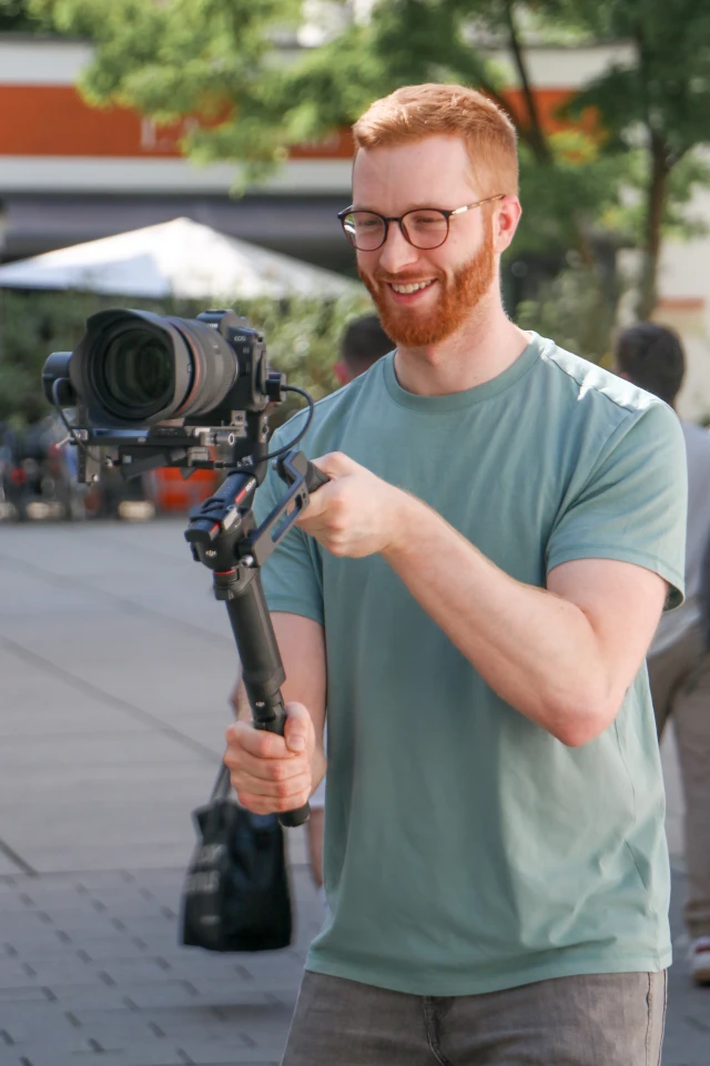 Ein Foto zeigt einen jungen Mann mit roten Haaren und Bart, der eine Brille und ein hellgrünes T-Shirt trägt und eine professionelle Kamera bedient, die auf einem stabilisierenden Gimbal montiert ist. Er lächelt, während er sich auf die Aufnahme der Bilder konzentriert. Die Szene spielt sich im Freien in einem gut beleuchteten Bereich ab, mit unscharfen Bäumen und Gebäuden im Hintergrund, die auf eine städtische Umgebung hindeuten. Der Mann scheint Spaß an seiner Aufgabe zu haben, und der Aufbau deutet auf eine professionelle oder semiprofessionelle Videoaufnahme hin.