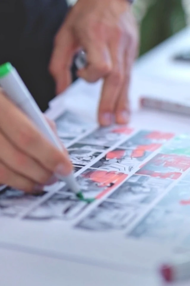 Eine Nahaufnahme zeigt die Hände einer Person bei der Arbeit an einem Comicstrip oder Storyboard. Die Person verwendet einen grünen Marker, um Teile der Schwarz-Weiß-Bilder zu färben oder hervorzuheben, auf denen verschiedene Tafeln mit Figuren und Szenen zu sehen sind. Der Fokus liegt auf den Händen und dem Kunstwerk, wobei der Hintergrund leicht verschwommen ist, was auf eine kreative und konzentrierte Tätigkeit hinweist. Die Szene deutet auf einen Künstler oder Illustrator hin, der gerade dabei ist, Details oder Anmerkungen zu seinem Werk hinzuzufügen.