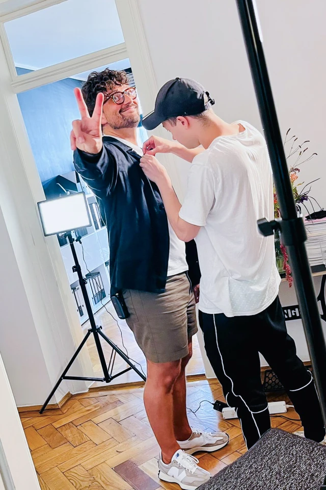 Ein Foto zeigt einen Mann, der in einem hell erleuchteten Innenraum steht, lächelt und mit seiner linken Hand ein Friedenszeichen zeigt. Er hat lockiges Haar und trägt eine Brille, eine dunkle Jacke, khakifarbene Shorts und weiße Turnschuhe. Eine zweite Person, die ein weißes T-Shirt, eine schwarze Hose und eine schwarze Kappe trägt, justiert gerade ein Mikrofon oder eine andere Ausrüstung an der Jacke des ersten Mannes. Im Hintergrund sind professionelle Beleuchtungsgeräte und Regale mit Büchern und Ordnern zu sehen, was auf ein Studio oder ein Büro schließen lässt. Die Gesamtstimmung ist heiter und professionell, wobei der erste Mann direkt in die Kamera schaut, während die zweite Person sich auf ihre Aufgabe konzentriert.