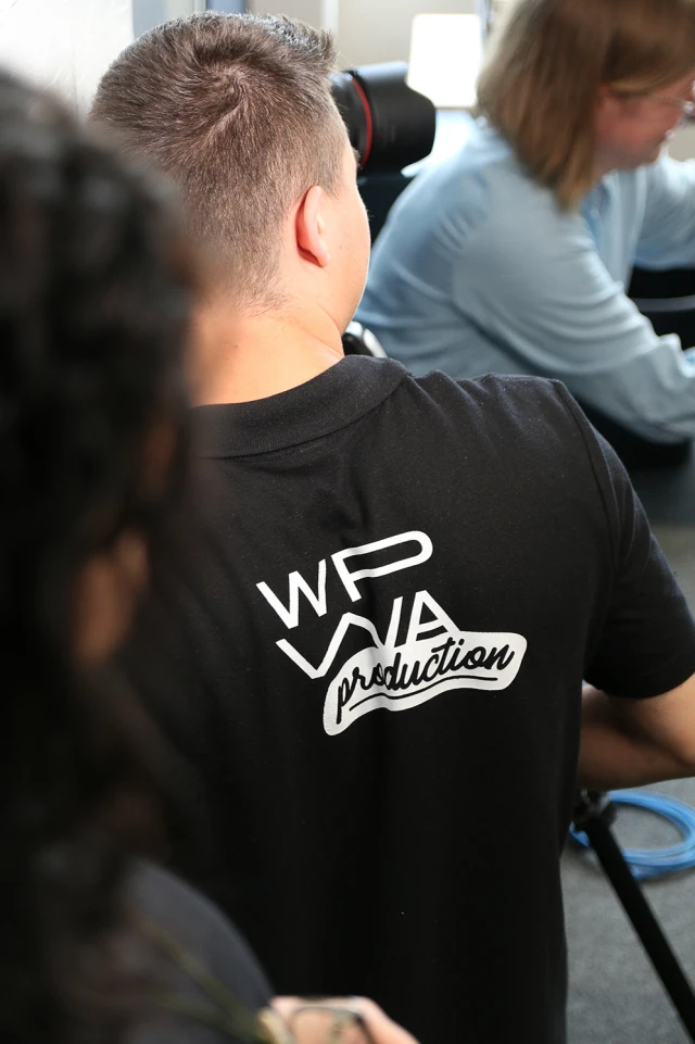 Auf dem Foto ist der Rücken einer Person zu sehen, die ein schwarzes Polohemd mit einem weißen Logo trägt. Die Person blickt von der Kamera weg, ihr kurzes Haar ist sichtbar. Im Hintergrund ist eine andere Person in einem hellblauen Hemd leicht unscharf zu erkennen, die sich auf eine Aufgabe oder Ausrüstung konzentriert. Die Umgebung scheint ein Innenraum zu sein, wahrscheinlich ein Studio oder eine Produktionsumgebung, in der ein Kameraobjektiv und andere Geräte teilweise sichtbar sind.