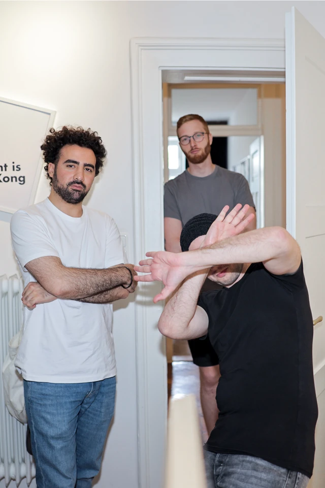 Ein Foto zeigt drei Männer in einem Flur. Der Mann auf der linken Seite hat lockiges Haar, einen Bart und trägt ein weißes T-Shirt mit verschränkten Armen. Der Mann in der Mitte, der eine Brille und einen Bart trägt, steht in der Tür und trägt ein graues T-Shirt. Der Mann auf der rechten Seite trägt eine schwarze Mütze und ein schwarzes T-Shirt, seine Arme sind erhoben und verdecken teilweise sein Gesicht, als ob sie die Kamera abhalten würden. Der Flur ist gut beleuchtet, und ein Teil eines gerahmten Bildes an der Wand ist sichtbar.