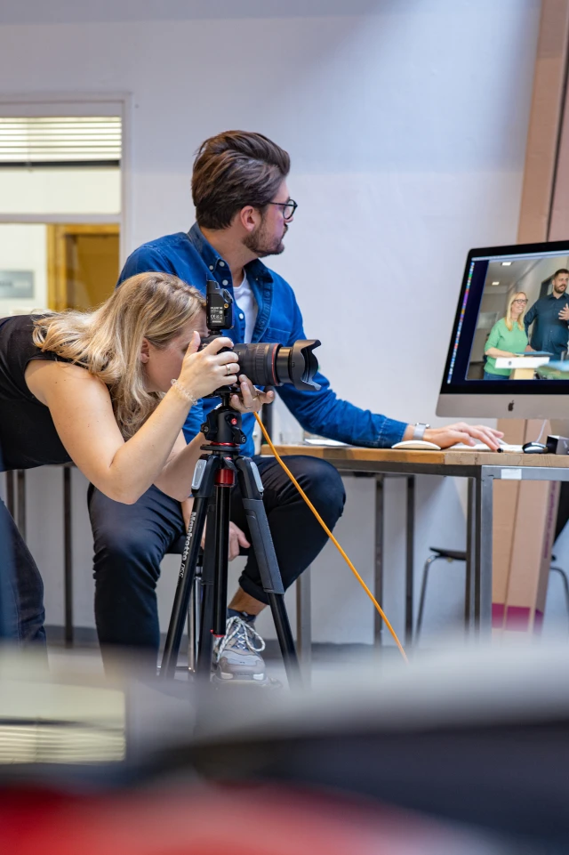 Ein Foto fängt einen Moment hinter den Kulissen in einer Studioumgebung ein. Eine Frau lehnt sich nach vorne, blickt durch den Sucher einer auf einem Stativ montierten Kamera und konzentriert sich ganz auf ihr Motiv. Sie hat blondes Haar und trägt ein schwarzes, ärmelloses Oberteil. Neben ihr sitzt ein Mann mit dunklem Haar, Brille und Bart und schaut auf einen Computerbildschirm. Er trägt ein blaues Jeanshemd und eine schwarze Hose. Auf dem Monitor ist ein Bild von zwei Personen, einem Mann und einer Frau, zu sehen, die sich anscheinend in einer professionellen Umgebung befinden. Die Szene ist gut beleuchtet, und verschiedene Studiogeräte und Möbel sind zu sehen, was zu der geschäftigen Atmosphäre beiträgt.
