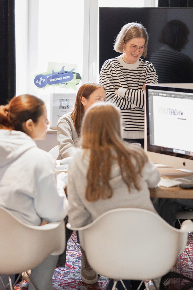 Eine Fotografie zeigt eine Gruppe junger Frauen, die in einem hellen, gut beleuchteten Raum zusammenarbeiten. Im Vordergrund sitzen drei Frauen an einem Tisch und konzentrieren sich auf einen großen Computermonitor, der ein Design oder einen Text anzeigt. Sie tragen legere Kleidung, darunter Kapuzenpullis. Im Hintergrund steht eine Frau mit kurzen blonden Haaren und Brille, gekleidet in einen gestreiften Pullover, lächelnd mit verschränkten Armen und beaufsichtigt die Aktivitäten. Die Umgebung scheint ein gemeinschaftlicher Arbeitsbereich zu sein, mit einem großen Fenster, das natürliches Licht hereinlässt, und einem dekorativen Teppich auf dem Boden. Im Hintergrund sind verschiedene Schilder und Materialien zu sehen, die auf eine kreative und engagierte Umgebung hindeuten.
