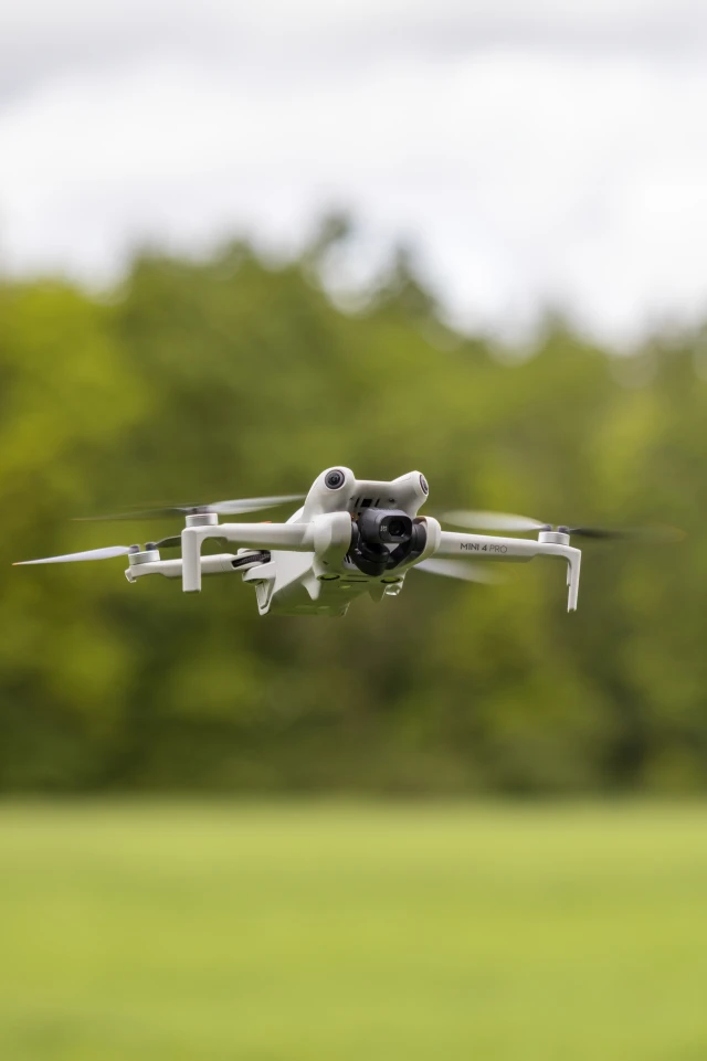 A photograph captures a close-up view of a small, white drone in flight. The drone is seen hovering in mid-air, with its four propellers visibly spinning. The background is an outdoor setting, featuring a lush, green, blurred backdrop of trees and grass, indicating a natural environment. The image is well-focused on the drone, highlighting its design and features, while the surroundings provide a soft, contrasting context.