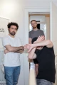 A photograph shows three men in a hallway. The man on the left has curly hair, a beard, and is wearing a white t-shirt with his arms crossed. The man in the middle, who has glasses and a beard, is standing in the doorway, wearing a gray t-shirt. The man on the right is wearing a black beanie and black t-shirt, with his arms raised, partially covering his face as if to block the camera. The hallway is well-lit, and a portion of a framed picture on the wall is visible.