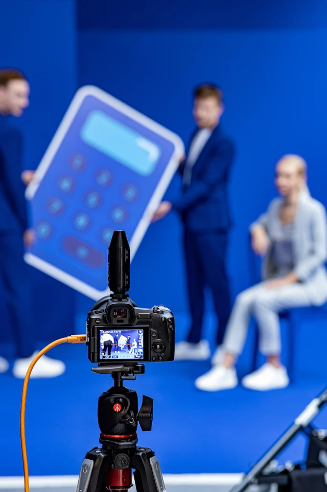 Ein Foto zeigt einen Aufbau hinter den Kulissen eines Fotoshootings. Im Vordergrund ist eine Kamera auf einem Stativ zu sehen, die die Szene einfängt. Auf dem Kamerabildschirm ist eine Live-Ansicht von zwei Personen im Hintergrund zu sehen, die ein großes, überdimensionales Modell eines blauen Taschenrechners halten. Die Personen sind leicht unscharf, tragen blaue Anzüge und stehen vor einem einfarbig blauen Hintergrund. Eine weitere Person, die helle Kleidung trägt, sitzt rechts, ebenfalls leicht unscharf. Die gesamte Szene hat ein professionelles und lebhaftes blaues Thema.