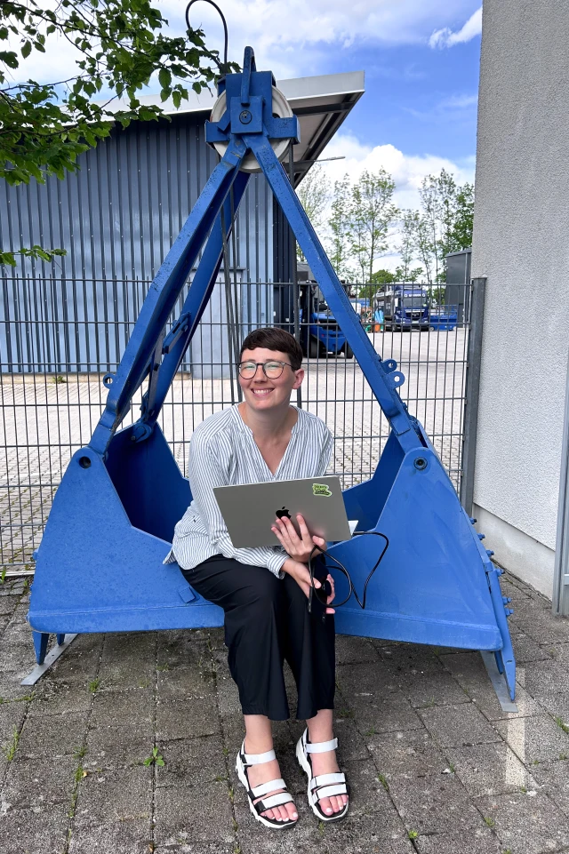 A photograph shows a woman sitting outside on a large, industrial blue metal grab bucket, typically used for heavy lifting or construction purposes. She has short dark hair and is wearing glasses, a striped white blouse, black pants, and white sandals. She is holding an open laptop on her lap and smiling, looking slightly to the side. The setting appears to be an industrial area with a fence, a blue building, and various equipment in the background. The weather is clear with a blue sky and some clouds visible, and there are trees in the background, indicating a pleasant day.
