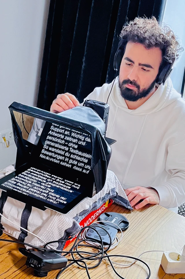 A photograph captures a man sitting at a wooden table, wearing a white hoodie and black headphones. He has curly hair and a beard, and he is focused on a teleprompter setup in front of him. The teleprompter screen displays white text on a black background, partially visible. The teleprompter is placed on top of a pile of books and covered with a cloth. Various cables and a camera lens cap are scattered on the table. The background features a dark curtain, adding contrast to the well-lit workspace.