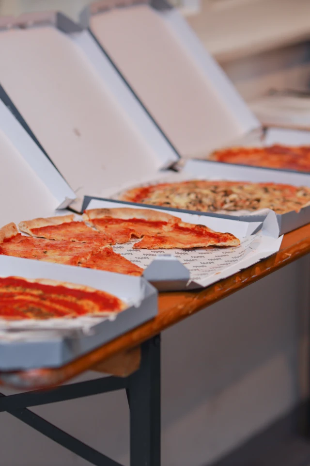 A photograph shows an array of pizzas in open boxes laid out on a table. There are four pizzas visible, each with a different topping. The pizzas are partially sliced, with some slices already missing. The boxes are slightly ajar, revealing the pizzas inside. The table is a simple wooden structure, and the background is blurred, suggesting an indoor setting. The overall scene suggests a casual meal, possibly in a communal or office environment.
