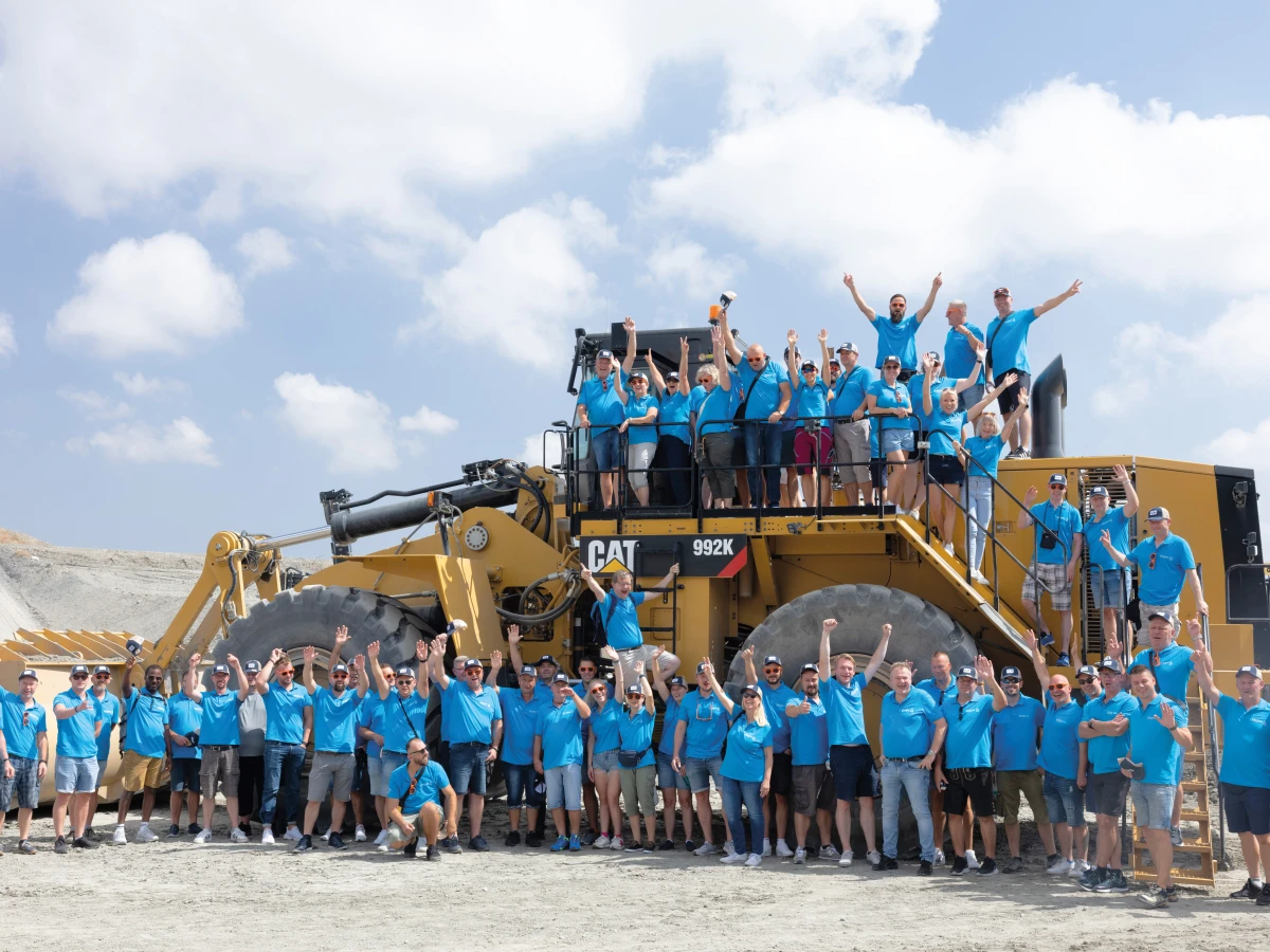 Auf einem Outdoor-Gelände versammelt sich ein großes Team vor und auf einer imposanten Baumaschine, während die Mitarbeitenden jubelnd ihre Begeisterung zeigen. Dieses Imagefoto steht für starken Teamgeist und technisches Interesse und wurde speziell für den Einsatz auf verschiedenen Social-Media-Kanälen sowie der Unternehmenswebsite erstellt.