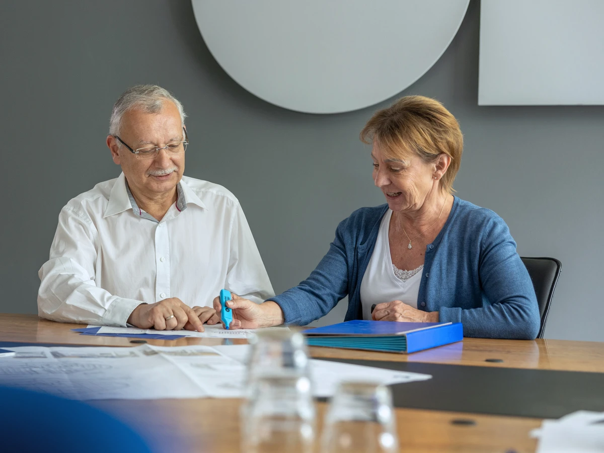 Ein Mann im weißen Hemd führt ein Gespräch mit seiner Kollegin am Konferenztisch. Der Blick beider Mitarbeiter richtet sich auf den Tisch, auf dem unternehmensspezifische Dokumente liegen. Dieses Imagefoto zeigt die professionelle und freundliche Zusammenarbeit, perfekt geeignet für die Darstellung der Dienstleistungen auf der Website und in sozialen Medien.