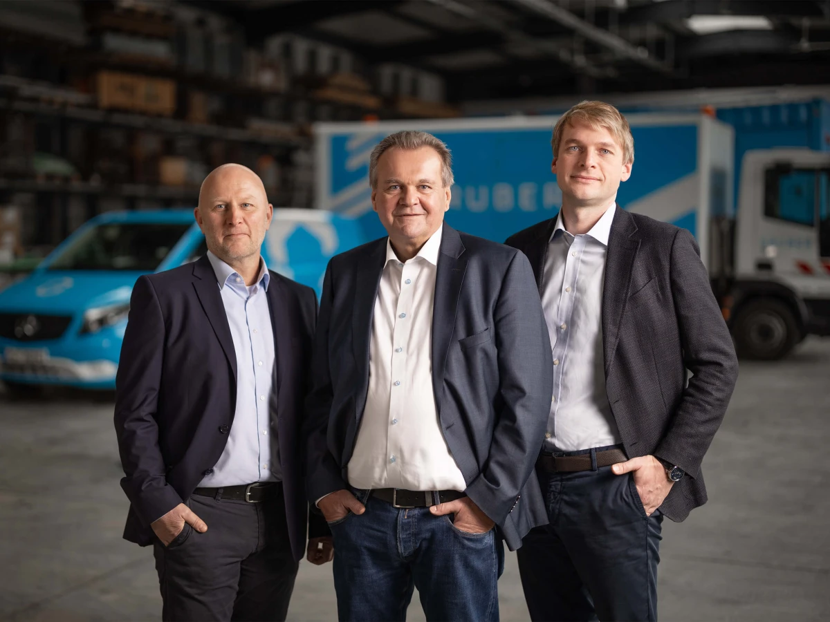 In a modern warehouse, three executives pose in front of company vehicles in a professional setting. This image was created for various corporate channels and LinkedIn profiles.