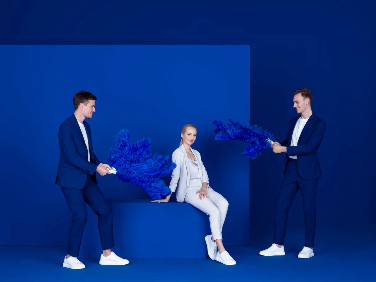 In einem professionellen Studio-Setting posiert eine Frau in einem weißen Outfit, während zwei Männer in blauen Anzügen mit blauen Federn für eine elegante Ausleuchtung sorgen. Das sorgfältig konzipierte Bühnenbild, das Casting der Models und das durchdachte Storyboard betonen die dramaturgische Inszenierung der Dienstleistungen des Kunden.