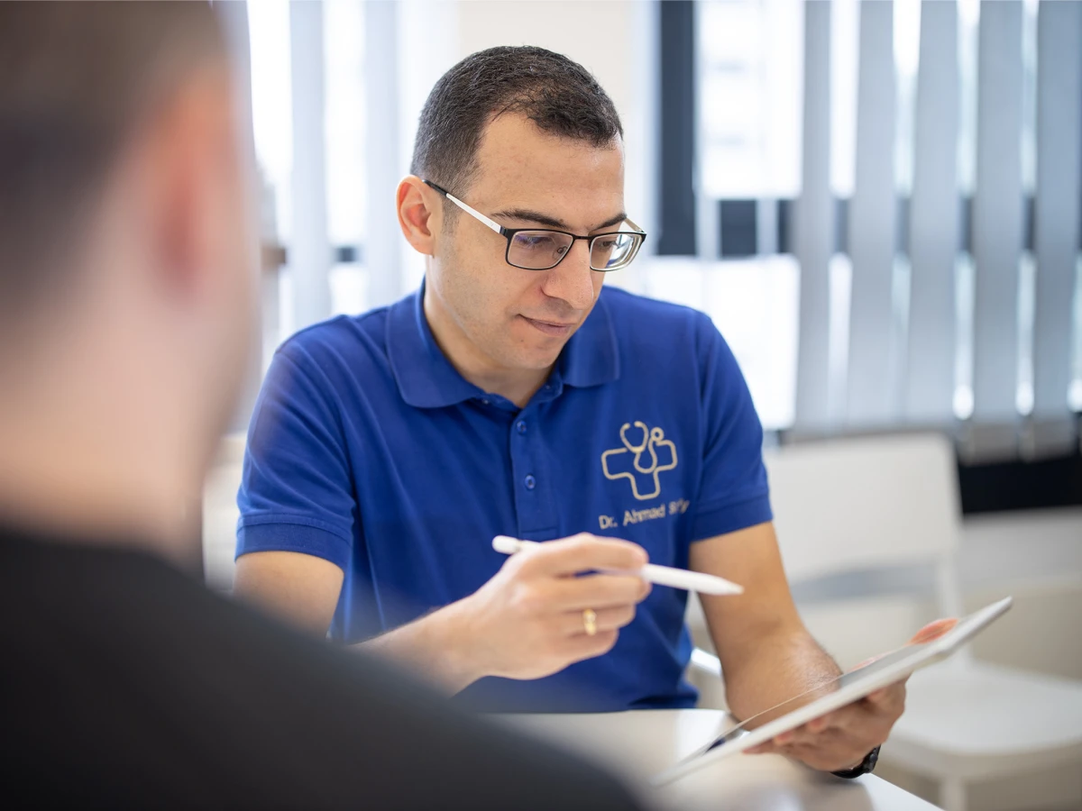 Ein Arzt im blauen Poloshirt führt ein Gespräch und macht Notizen auf einem Tablet. Dieses Imagefoto zeigt die professionelle und patientenorientierte Betreuung in der Arztpraxis, perfekt geeignet für die Darstellung der Dienstleistungen auf der Website und in sozialen Medien.