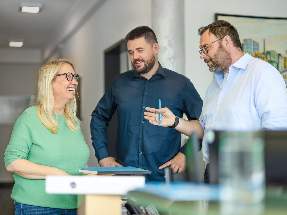 Ein Team aus zwei Männern und einer Frau ist um einen Tisch versammelt. Sie blicken zueinander und unterhalten sich freundlich. Dieses Imagefoto zeigt das positive Arbeitsklima im Unternehmen, perfekt geeignet fürbass Employer Management auf der Website oder in den sozialen Medien.