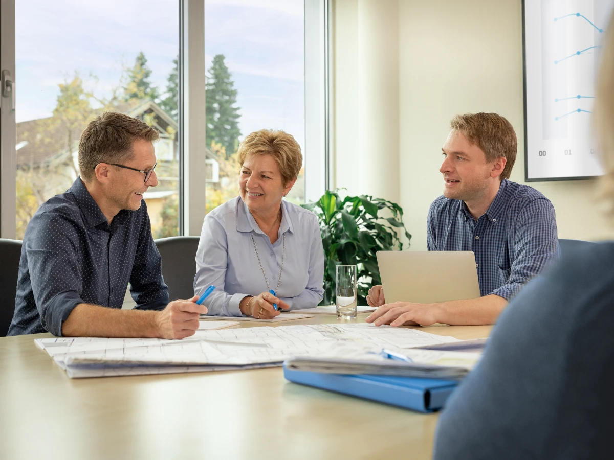 Ein Team hält ein Meeting am Konferenztisch, während technische Baupläne vor ihnen liegen. Die Mitarbeitenden blicken einander freundlich an und tauschen sich engagiert aus. Dieses Imagefoto veranschaulicht die professionelle und kollegiale Zusammenarbeit und eignet sich ideal zur Präsentation der Dienstleistungen auf der Website und in sozialen Medien.