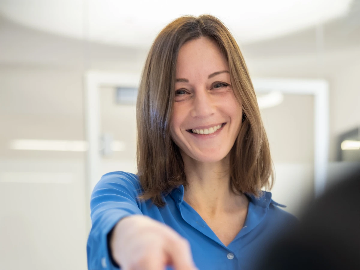 A smiling receptionist hands over a card across the desk. This image conveys the welcoming and professional atmosphere of the medical practice, ideal for introducing the team and for use across various channels.